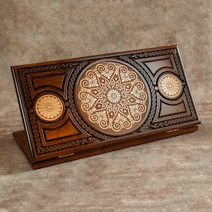 Könnte beinhalten: Ein Holzspielbrett mit aufwendigen Schnitzereien und einem goldfarbenen Intarsienmuster. Das Brett ist geschlossen und hat einen Klappdeckel.