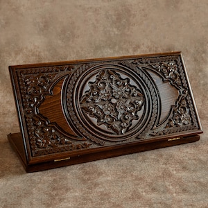 Puede incluir: Un tablero de backgammon de madera marrón oscuro con intrincadas tallas florales. El tablero presenta un diseño circular central y patrones detallados en los bordes. El tablero está abierto, revelando la superficie de juego y las bisagras de latón.