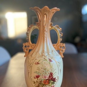 May include: A vintage porcelain vase with ornate gold accents and a floral design. The vase has two handles and a flared top.