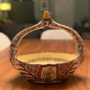 May include: A ceramic bowl with a handle shaped like a bird. The bowl is decorated with pink, green, yellow, and white floral patterns.