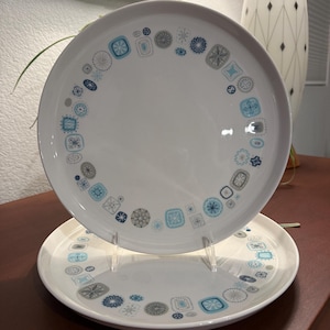May include: Two white ceramic plates with a repeating pattern of blue, gray, and teal geometric shapes. The plates are round and have a glossy finish. The design is arranged around the rim of the plates. The plates are displayed on a brown surface.