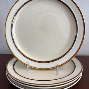 May include: A stack of four ceramic plates. The plates are off-white with a speckled texture and a brown border. The plates are stacked on a dark brown surface.