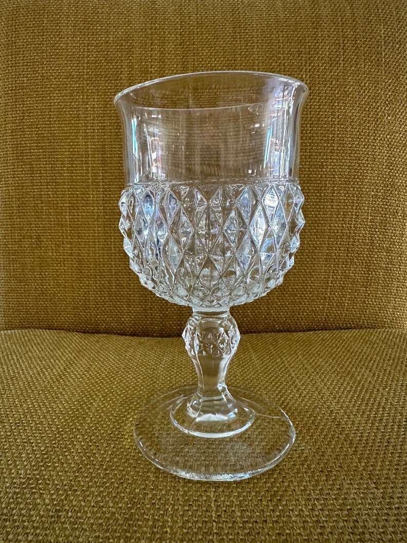 May include: A clear glass goblet with a diamond-shaped pattern. The goblet has a tall, slender stem and a wide, round base.