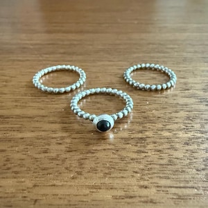 May include: Three silver rings with beaded bands are displayed on a wooden surface. One ring features a dark, possibly black, central stone. The other two rings are simple beaded bands. The rings are likely for the fingers.