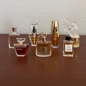 May include: A collection of six miniature perfume bottles of varying shapes and sizes. The bottles contain liquids in shades of clear, amber, and deep red. Labels include Poeme, True Love, and Chanel Gardenia, with one bottle featuring a clear glass bird topper.