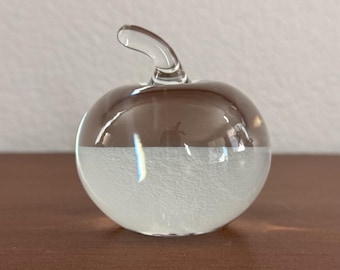 May include: A clear glass apple sculpture with a brown top and a white, textured bottom. The apple has a curved stem and sits on a brown surface. The sculpture is a decorative item.