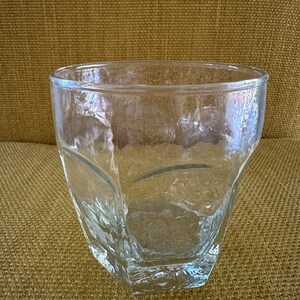 May include: A clear glass tumbler with a textured, faceted design. The tumbler is empty and sitting on a brown textured surface.