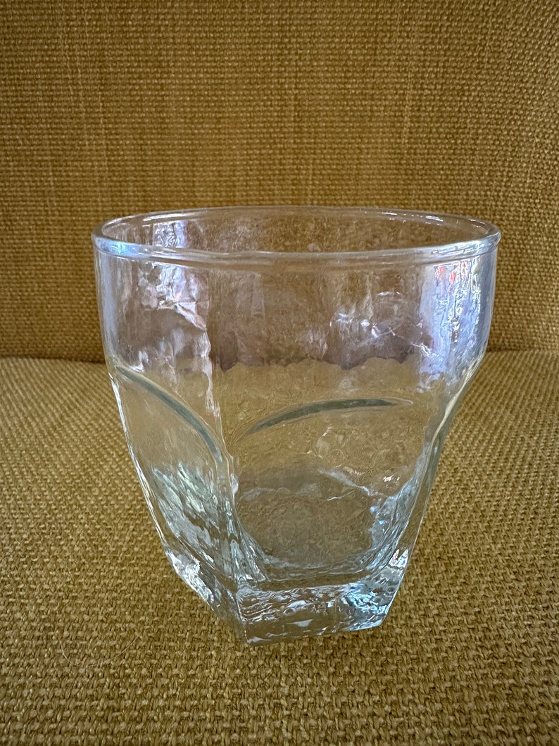 May include: A clear glass tumbler with a textured, faceted design. The tumbler is empty and sitting on a brown textured surface.