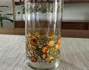 May include: A clear glass vase with a floral pattern featuring yellow, red, and green fruits and vegetables. The vase has a gold rim and a green leafy design around the top.