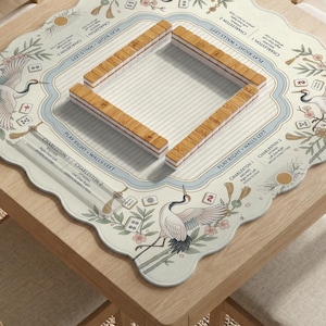 May include: A square mahjong mat with scalloped edges featuring illustrations of white cranes, bamboo, and cherry blossoms. The mat includes printed text for Charleston rules and play directions, with a central square of wooden mahjong tiles.
