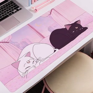 Könnte beinhalten: Ein rechteckiges rosa Mauspad mit einer Cartoon-Illustration von zwei Katzen, einer weißen und einer schwarzen, die auf einer rosa Oberfläche ruhen. Der Hintergrund zeigt eine Stadtlandschaft und einen Mond. Ein Laptop und rosa Zubehör sind sichtbar.