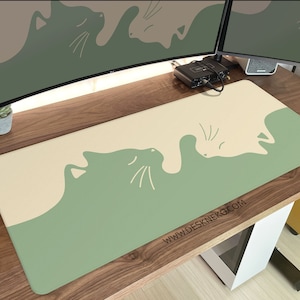 May include: A rectangular desk mat featuring a cat design in sage green and cream colors. The design shows two stylized cat silhouettes facing each other. The website address www.deskneko.com is printed on the mat.