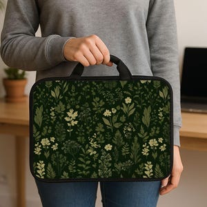 Può includere: Una custodia nera per laptop con manico, con sfondo verde scuro e un motivo di illustrazioni botaniche verde chiaro e bianche. La custodia è tenuta da una persona.