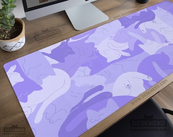 May include: A rectangular desk mat featuring a pattern of stylized cats in shades of purple and white. The mat is placed on a wooden desk, with a computer monitor, a potted plant, and a black cup visible in the background. The text "DeskNeko" is visible on the mat.