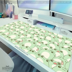 May include: A large desk mat featuring a repeating pattern of cartoon frogs in light green and cream colors. The frogs have their tongues sticking out. The website address www.DESKNEKO.COM is printed on the mat. The mat is on a white desk.