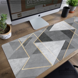 May include: A gray desk mat with a geometric design of triangles and gold lines. The mat is rectangular and covers a wooden desk surface. A computer monitor, a small plant, and a black cup are also on the desk. The website address www.DESKNEKO.COM is printed on the mat.