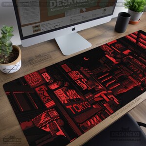 Op de afbeelding: Zwart en rode bureau mat met een gepixeld stadsbeeld van Tokio, inclusief Japanse tekst en het woord "TOKYO". De mat heeft een maan en de tekst "DeskNeko". De rechthoekige mat ligt op een houten bureau.