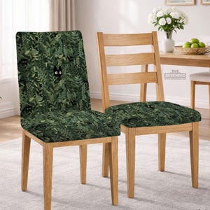 May include: Two wooden chairs. One chair has a seat and back covered in a dark green fabric with a pattern of black cat faces among green leaves. The other chair is natural wood with a green cat-themed seat cover. The chairs are in a dining room setting.