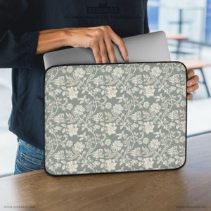 Pode incluir: Uma capa para laptop com um padrão floral. A capa é de cor verde suave com um design repetido de flores brancas e vinhas. Possui um zíper preto e contém um laptop prateado. O texto "DESKNEKO" é visível.