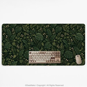 K&ouml;nnte beinhalten: Eine rechteckige Schreibtischunterlage mit einem dichten, dunkelgr&uuml;nen botanischen Muster aus Monstera-Bl&auml;ttern, Farnen und kleinen wei&szlig;en Bl&uuml;ten. Eine beige mechanische Tastatur und eine passende beige Computermaus liegen auf der Unterlage.