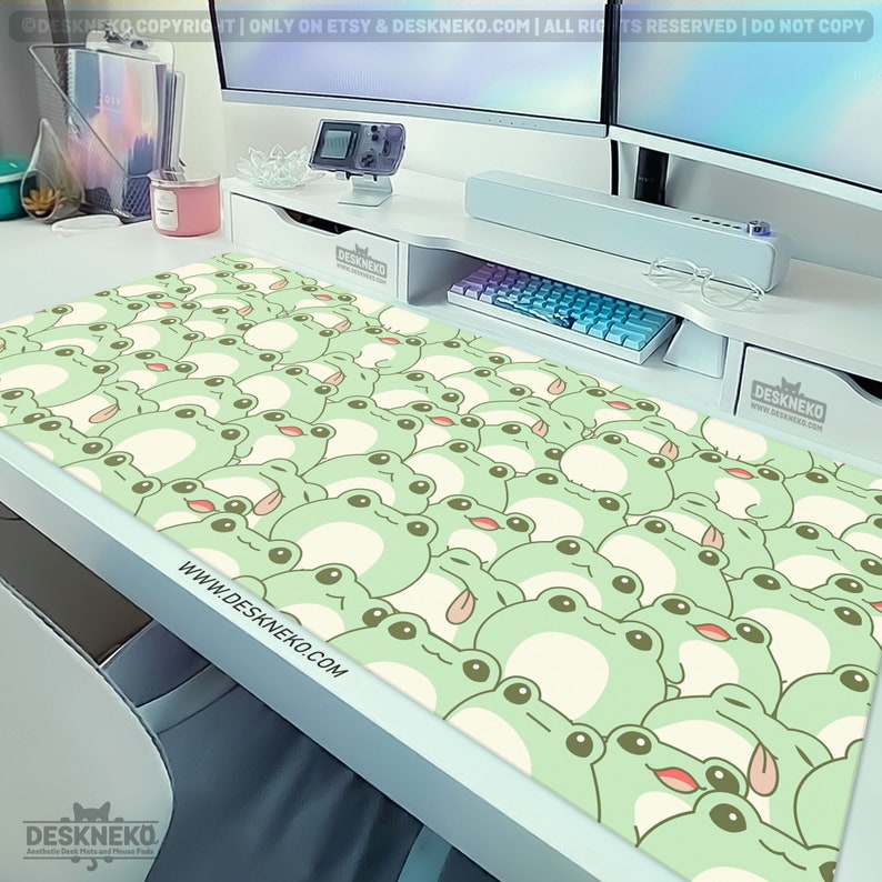 May include: A large desk mat featuring a repeating pattern of cartoon frogs in light green and cream colors. The frogs have their tongues sticking out. The website address www.DESKNEKO.COM is printed on the mat. The mat is on a white desk.