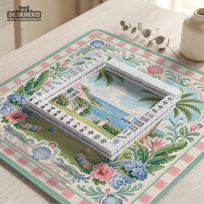 May include: A white Mahjong set with red and black characters is arranged on a decorative mat. The mat features a coastal scene with a house, ocean, and sailboat, surrounded by floral and seashell motifs. The mat has a pink and green border.