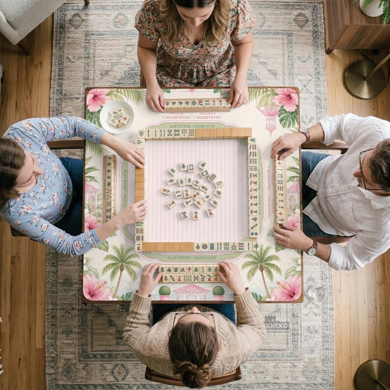 May include: A square mahjong game table featuring a light-colored surface with a tropical pattern of pink hibiscus flowers and green palm trees. The table includes a central playing area with scattered tiles and four racks holding rows of mahjong tiles.