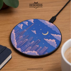 Puede incluir: Cargador inalámbrico redondo con un diseño de paisaje urbano que presenta una torre, edificios, nubes y una luna creciente contra un cielo azul. El cargador tiene un borde y un cable negros. La marca "Deskneko" está en la parte superior.