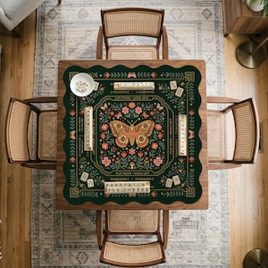 May include: A square dark green mahjong mat with scalloped edges, featuring a central butterfly and floral illustration. The mat includes printed game instructions, directional text, and scattered mahjong tiles. A small white bowl with tiles sits on the corner.