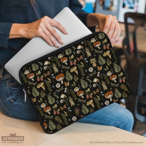 May include: Black laptop sleeve with a forest-themed design, including mushrooms, dragonflies, berries, and ferns. The sleeve has a black zip. The laptop sleeve is being held by a person. The sleeve is designed to hold a laptop.