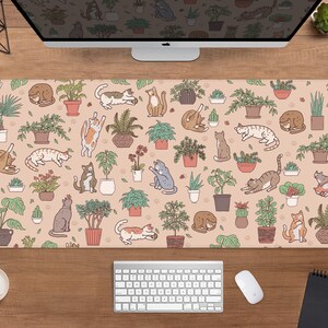 Puede incluir: Alfombrilla de escritorio rectangular con un patr&oacute;n repetido de gatos de dibujos animados y plantas en macetas sobre un fondo beige. Los gatos est&aacute;n en varias poses, y las plantas est&aacute;n en macetas marrones. Se ven una computadora, un teclado y un rat&oacute;n.