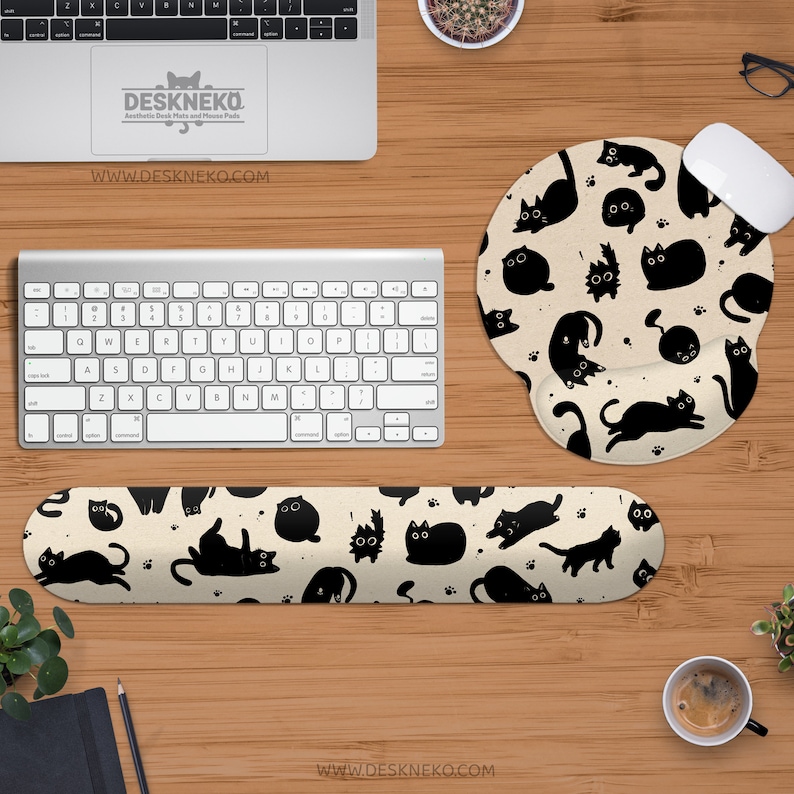 Puede incluir: Una configuraci&oacute;n de escritorio con teclado, rat&oacute;n y accesorios. La alfombrilla de rat&oacute;n y el reposamu&ntilde;ecas tienen un fondo beige con un patr&oacute;n de ilustraciones de gatos negros. El teclado es plateado y el rat&oacute;n es blanco.