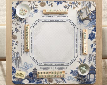 Op de afbeelding: Een vierkante Mahjong-mat met een blauw-wit bloemmotief en pagode-illustraties. De mat bevat gedrukte spelinstructies, vier houten tegelhouders, twee kleine schaaltjes met tegels en markers, en een klein kaartje met een merknaam.