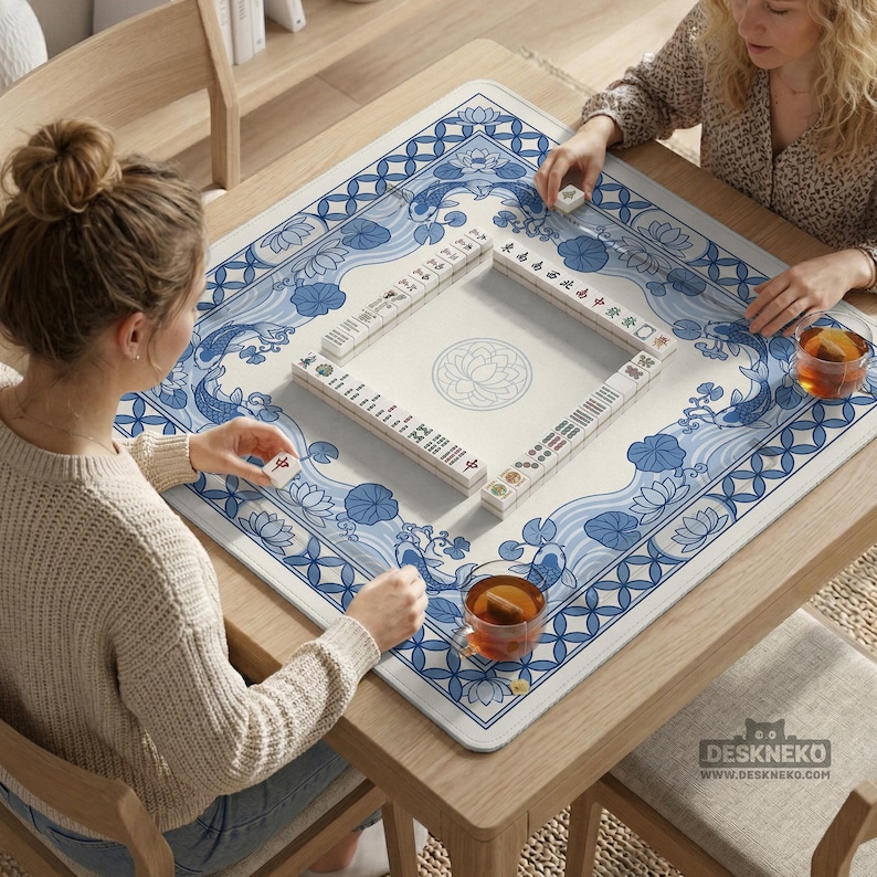 May include: A square wooden table with a blue and white patterned Mahjong mat. The mat features a floral and wave design. Two people are playing Mahjong, with tiles and tea cups visible. The website "DESKNEKO" is printed on the mat.