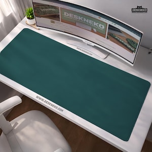 May include: A teal desk mat with rounded corners, covering most of a white desk. The mat has the website address www.deskneko.com printed on it. A curved computer monitor and a small plant are visible in the background.