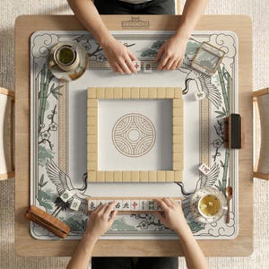 May include: Overhead view of a Mahjong game in progress. The beige tiles are arranged on a cream-colored mat with crane and bamboo designs. The wooden table has a logo that reads DESKNEKO. Two people are playing the game, with tea cups and accessories.