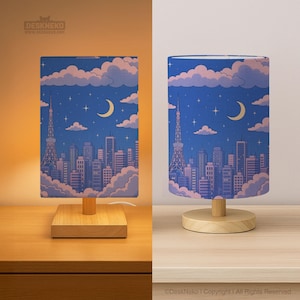 May include: Two table lamps with rectangular shades. The shades display a cityscape design with a blue backdrop, a crescent moon, clouds, and a tower. The lamps have a wooden base and a light bulb inside.