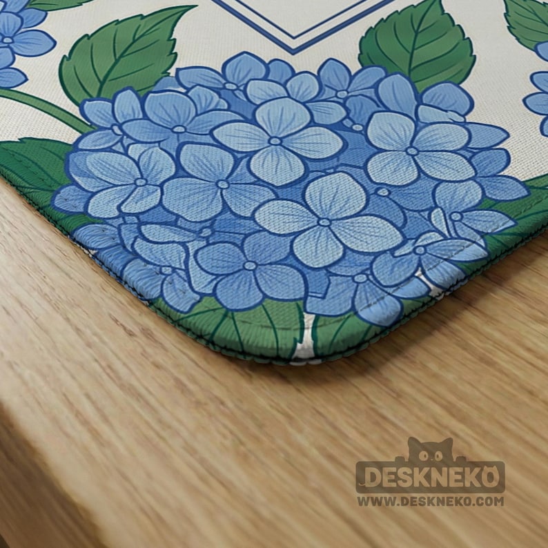 May include: A corner of a desk mat featuring a blue hydrangea floral design. The mat has a white background with green leaves and a blue border. The mat is on a wooden surface. The logo for DESKNEKO is visible in the bottom right corner.