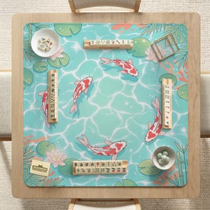 May include: A square mahjong mat featuring a light blue water surface pattern with koi fish, lily pads, and lotus flowers. The mat includes four wooden tile racks, two small white bowls containing mahjong tiles, and a small card labeled DESKNEKO.