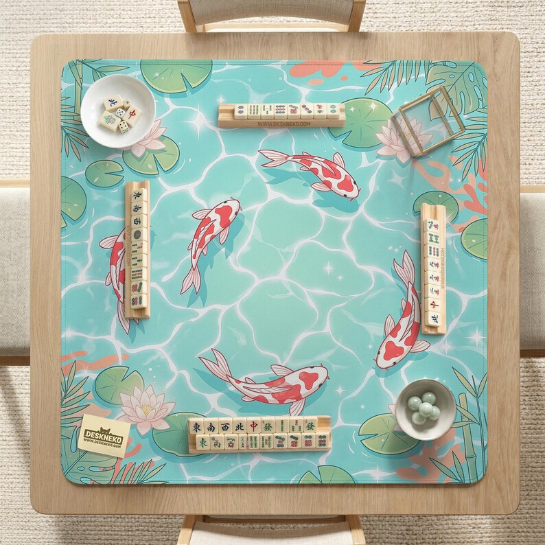 May include: A square mahjong mat featuring a light blue water surface pattern with koi fish, lily pads, and lotus flowers. The mat includes four wooden tile racks, two small white bowls containing mahjong tiles, and a small card labeled DESKNEKO.
