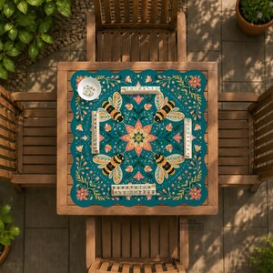 May include: A square game mat with a scalloped edge featuring a teal background, a central pink floral mandala, and four bees in the corners. The mat is decorated with botanical vines and flowers, and includes four wooden tile racks and a small white bowl.