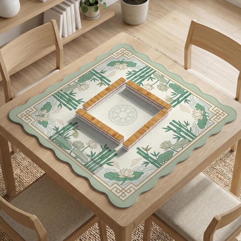 Puede incluir: Un tapete de mesa de mahjong cuadrado con un borde festoneado, con un fondo crema con dise&ntilde;os de bamb&uacute; y flores de loto verdes. La alfombra rodea un &aacute;rea cuadrada con un patr&oacute;n circular, y las fichas de mahjong de madera se colocan encima.