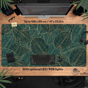 May include: A large rectangular desk mat with a teal and gold leaf pattern. The mat measures up to 120 x 60 cm. A black keyboard, mouse, and other accessories are on the mat. The text "With optional LED/RGB lights" is visible.