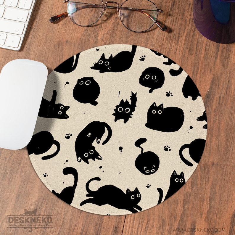 Puede incluir: Alfombrilla de rat&oacute;n redonda beige con un patr&oacute;n repetido de ilustraciones de gatos negros en varias poses. La alfombrilla tiene el texto "DESKNEKO" y "Aesthetic Desk Mats with Hand Paws". Se ve un rat&oacute;n y un teclado blancos.