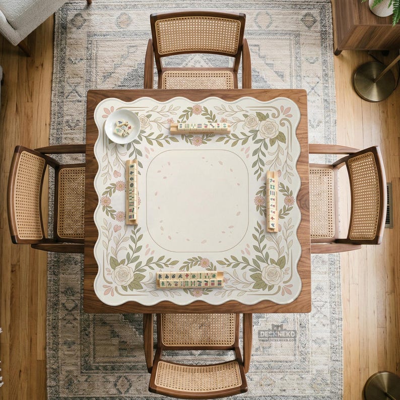 May include: A square wooden table with four cane-back chairs, featuring a scalloped-edge mat with a floral vine pattern in muted green and pink tones. The mat displays mahjong tiles arranged in racks and a small white bowl with loose tiles.
