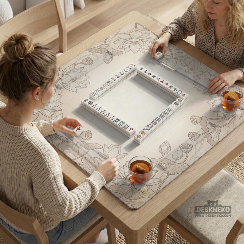 Puede incluir: Una mesa de mahjong de madera con un tapete de dise&ntilde;o floral. El tapete presenta un patr&oacute;n de flor de loto en gris y blanco. Las fichas de mahjong blancas est&aacute;n dispuestas en un cuadrado. Dos personas juegan al mahjong, cada una con una taza de t&eacute;.