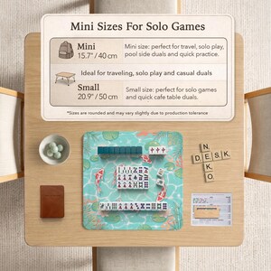 May include: A mahjong set on a light blue mat with koi fish and lily pad illustrations. The mat includes a chart titled Mini Sizes For Solo Games, listing 15.7 inch and 20.9 inch options. Wooden letter tiles spell Neko Desk, with a small bowl of marbles.