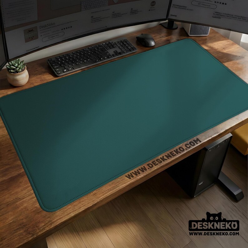 Op de afbeelding: Een grote, rechthoekige teal bureaumat met gestikte randen. De mat ligt op een houten bureau, met een toetsenbord, muis en monitor zichtbaar. Het websiteadres WWW.DESKNEKO.COM is op de mat gedrukt.
