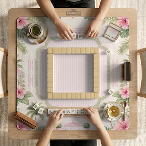 May include: A square mahjong mat featuring a pink and green tropical hibiscus floral pattern with palm trees and a gazebo. The mat includes printed Charleston rules and a central square of mahjong tiles, with two tea cups and a tile rack visible.