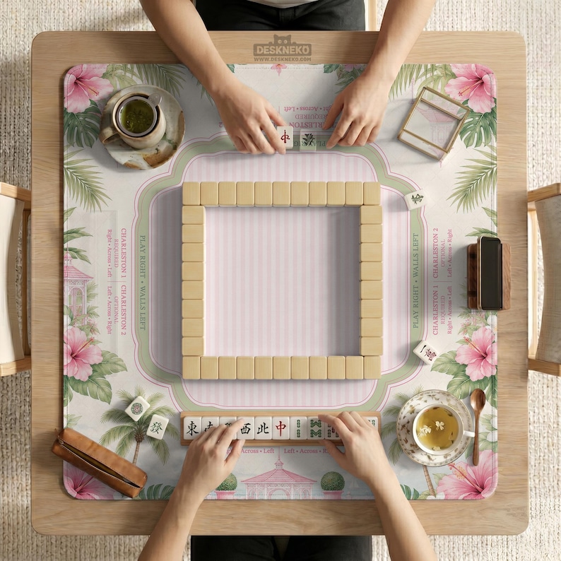 May include: A square mahjong mat featuring a pink and green tropical hibiscus floral pattern with palm trees and a gazebo. The mat includes printed Charleston rules and a central square of mahjong tiles, with two tea cups and a tile rack visible.